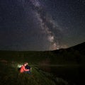 Two young people sit at lake on background of mighty mountains under the starry sky