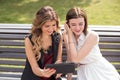 Two young girls sitting on a bench in a park watching the tablet and laughing Royalty Free Stock Photo