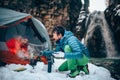 Two young professional male tourists are preparing food and hot drinks in the mountains near the river in winter.