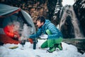 Two young professional male tourists are preparing food and hot drinks in the mountains near the river in winter.