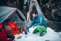 Two young professional male tourists are preparing food and hot drinks in the mountains near the river in winter.