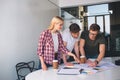 Two young serious men stand at table and work. One of them white on big piece of paper. Young blonde woman point on it Royalty Free Stock Photo