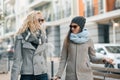 Two young smiling beautiful women in warm clothes walking down the city street with a travel suitcase, women laughing and talking Royalty Free Stock Photo