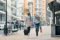 Two young smiling beautiful women in warm clothes walking down the city street with a travel suitcase, women laughing and talking Royalty Free Stock Photo