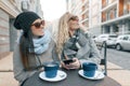 Two young smiling fashionable women in an outdoor cafe, drinking coffee, talking, laughing. Urban background Royalty Free Stock Photo