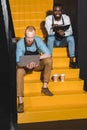 two young smiling owners of coffee shop in aprons with laptop and textbook sitting on stairs with paper cups of