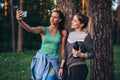 Two young sportive girlfriends wearing sportswear leaning against tree taking selfie with smartphone in forest