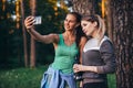 Two young sportive girlfriends wearing sportswear leaning against tree taking selfie with smartphone in forest