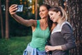 Two young sportive girlfriends wearing sportswear leaning against tree taking selfie with smartphone in forest