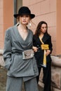 Two young stylish beautiful women fashion models are posing in street, wearing pantsuit, hat, having purse on waist.