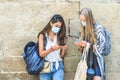 Two young tourists dressing casual and with backpacks and purses checking their mobile phones near a wall Royalty Free Stock Photo