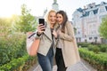 Two young women having fun, looking at the smartphone laughing, sunny autumn day, city background, golden hour