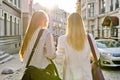 Two young women with laptop bags walking along street of sunset city, back view