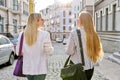 Two young women with laptop bags walking along street of sunset city, back view