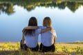 Two young women rear view hugging while sitting on the grass near the river in autumn. Friends leaned on each other in the park Royalty Free Stock Photo