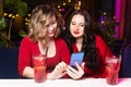 Two young women in red clothes drink cocktails and celebrate in a nightclub or bar. Girlfriends take selfies and smile