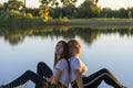 Two young women are resting in summer by the river. Blonde and brunette are back to back in nature. Friendship concept, soft focus Royalty Free Stock Photo