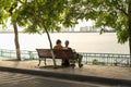 Two young women sitting on bench under big tree by the lake in city Royalty Free Stock Photo