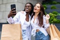 Summer sunny day. Two young women standing outdoors with shopping bags and make selfie on smartphone, looking on screen of phone Royalty Free Stock Photo