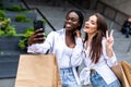 Summer sunny day. Two young women standing outdoors with shopping bags and make selfie on smartphone, looking on screen of phone Royalty Free Stock Photo