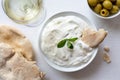 Tzatziki in white ceramic bowl with mint leaf garnish and a piece of pita bread next to pita bread, olives and a glass of white w