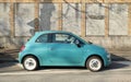 Light blue Fiat 500 hybrid with white rims. Side view with concrete and brick wall on background.