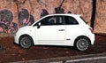 White Fiat 500 with small italian flag emblems at the roadside in front of a brick wall covered with graffiti. Side view.