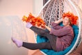 Ugly aged woman in a sweater with a wreath of yellow autumn maple leaves in a white wicker hanging chair in the room with curtains Royalty Free Stock Photo