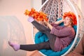 Ugly aged woman in a sweater with a wreath of yellow autumn maple leaves in a white wicker hanging chair in the room with curtains Royalty Free Stock Photo