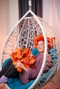 Ugly aged woman in a sweater with a wreath of yellow autumn maple leaves in a white wicker hanging chair in the room with curtains Royalty Free Stock Photo