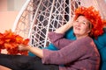 Ugly aged woman in a sweater with a wreath of yellow autumn maple leaves in a white wicker hanging chair in the room with curtains Royalty Free Stock Photo