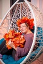 Ugly aged woman in a sweater with a wreath of yellow autumn maple leaves in a white wicker hanging chair in the room with curtains Royalty Free Stock Photo