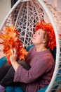 Ugly aged woman in a sweater with a wreath of yellow autumn maple leaves in a white wicker hanging chair in the room with curtains Royalty Free Stock Photo