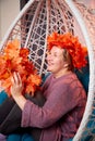 Ugly aged woman in a sweater with a wreath of yellow autumn maple leaves in a white wicker hanging chair in the room with curtains Royalty Free Stock Photo