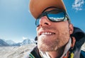 Ultra-wide lens angle portrait shot of high altitude mountain smiling unshaven happy hiker in baseball cap with snow peaks and