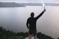 Ultras hooligan holding blue flare torch in hand, standing on top of rock mountain in evening with amazing view on river.