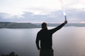 Ultras hooligan holding blue flare torch in hand, standing on top of rock mountain in evening with amazing view on river.