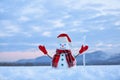 Unbelievable sunrise. Happy snowman in hat, scarf, red gloves with ice pikestaff is standing on the snow lawn. Mountains landscape