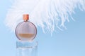 Unbranded round perfume bottle on glass podium and large white ostrich feather on blue background. Transparent glass perfume