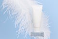 Unbranded white squeeze bottle cream tube on glass podium and large white ostrich feather on blue background. Lotion, balsam, hand