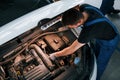 Under the hood. Man in work uniform repairs white automobile indoors. Conception of automobile service