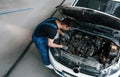 Under the hood. Man in work uniform repairs white automobile indoors. Conception of automobile service