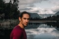 unfocused disheveled caucasian guy with beard calm and relaxed looking at camera at lonely cold water lake among forest