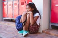 Unhappy african american schoolgirl sitting by lockers in school corridor with schoolbag Royalty Free Stock Photo