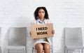 Unhappy African American woman holding I NEED WORK sign while waiting for employment interview at company lobby Royalty Free Stock Photo