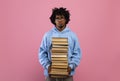 Unhappy black teen holding giant stack of books, looking at camera with bored face expression on pink background