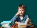 Unhappy boy of European appearance is sitting on the background of the school green board. Teen does not want to school. A stack