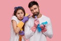 Unhappy displeased woman and man hold spray, sponges, dressed in white casual clothes, involved in cleaning, do house