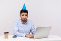 Unhappy employee guy with funny party cone on head sitting in office workplace, using laptop and looking depressed Royalty Free Stock Photo
