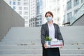 Unhappy fired masked woman is standing in the street with a box of personal items from the desktop. An unemployed
